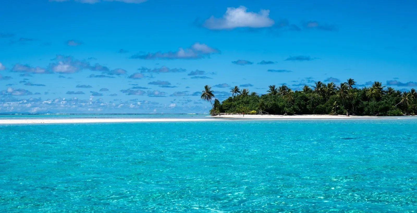 Clear turquoise lagoon water in front of a low palm islet and a white reef line