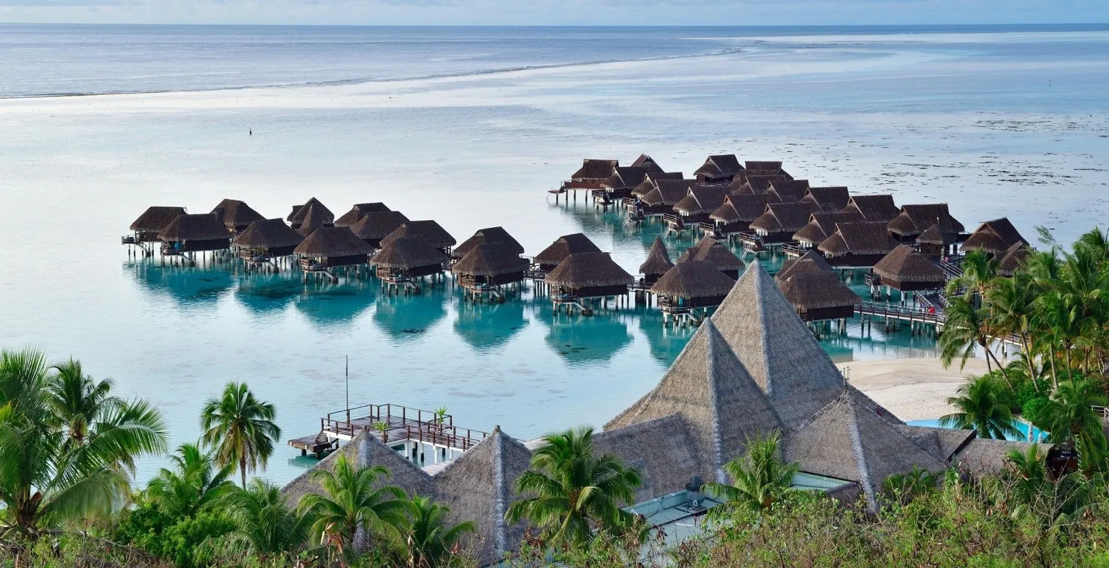 Overwater bungalows on a pale lagoon, seen past palm trees and thatched roofs